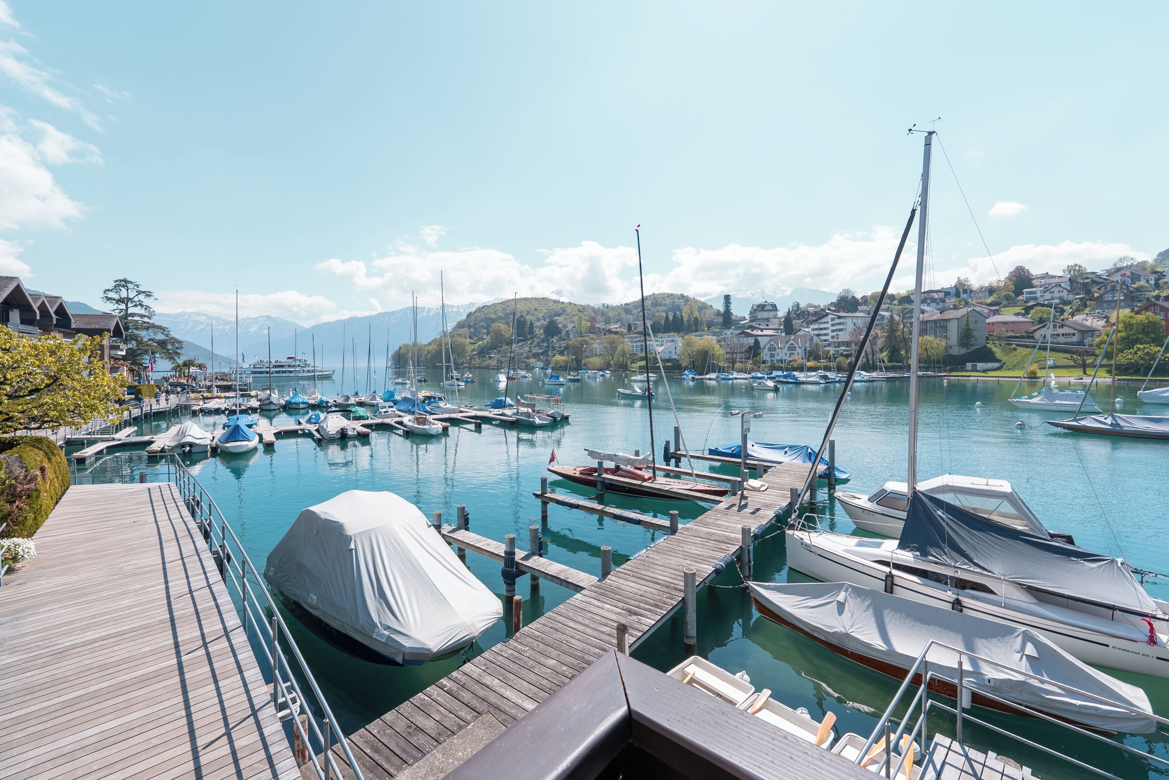 Un puerto deportivo con barcos amarrados, un muelle de madera y un fondo de casas y montañas.