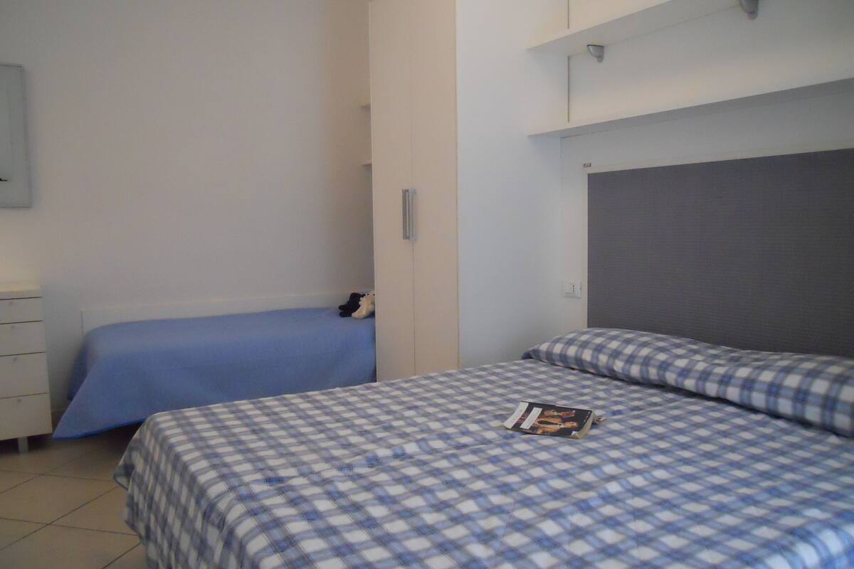 A bedroom with two beds, one with a blue bedspread and the other with a checkered bedspread. There is a wardrobe and a small table with a book on it.