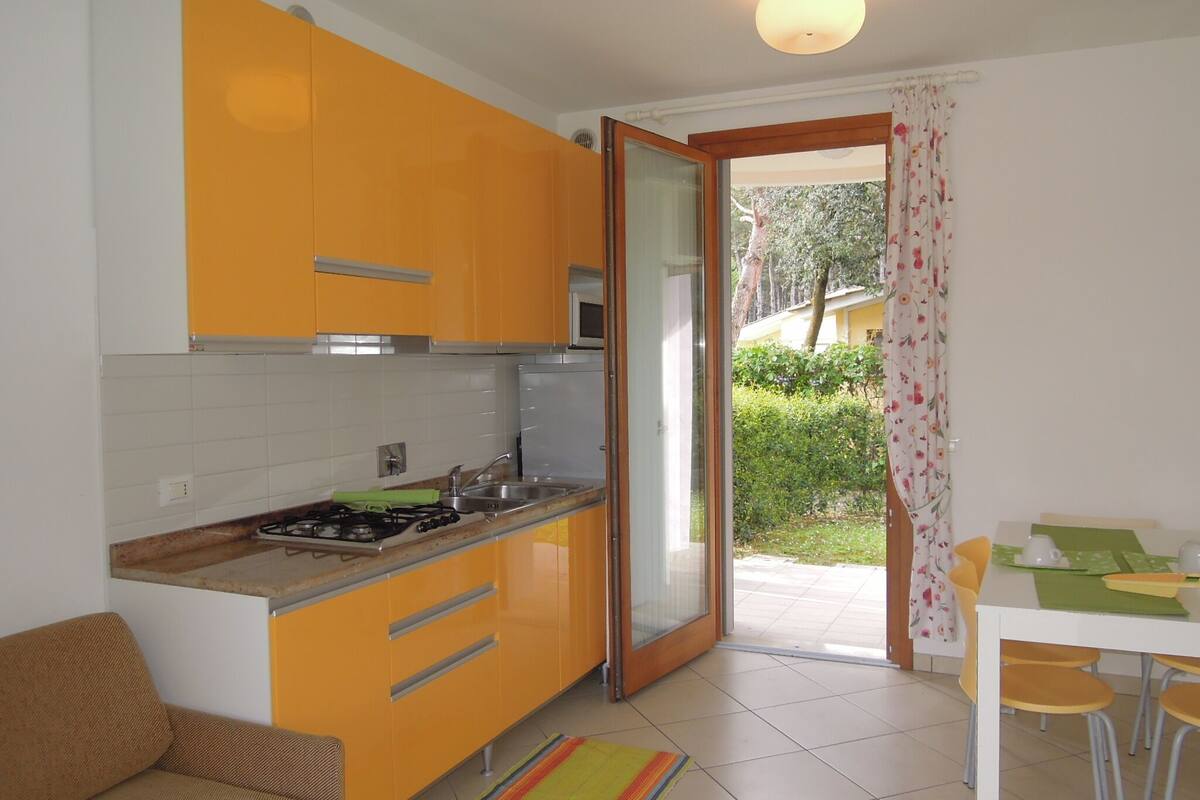 A compact kitchen with orange cabinets, a gas stove, and a microwave. There's a dining area with a table and chairs, and a sliding glass door leading to a garden.