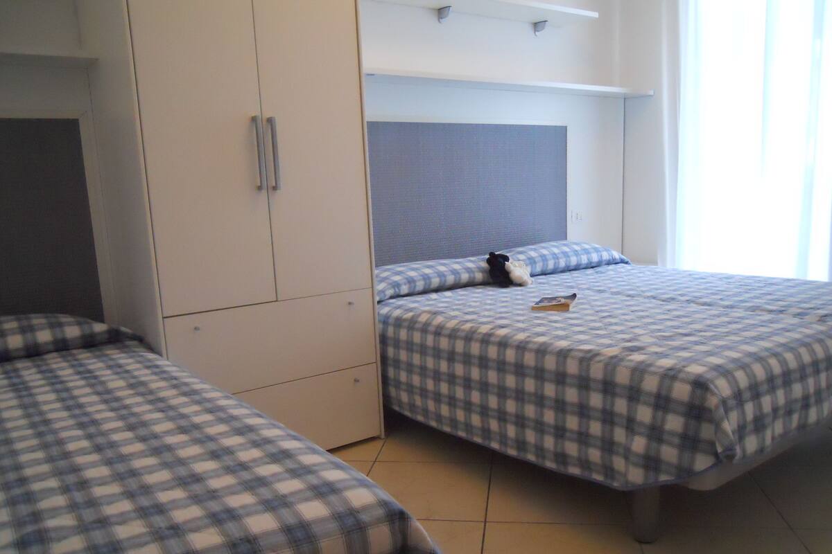 A room with two beds, each with a checkered bedspread. There is a wardrobe with a mirror and a small table with a book on it.