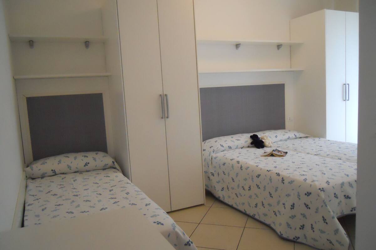 A bedroom with two beds, each with a headboard and a nightstand. There are wardrobes and a shelf above the beds.