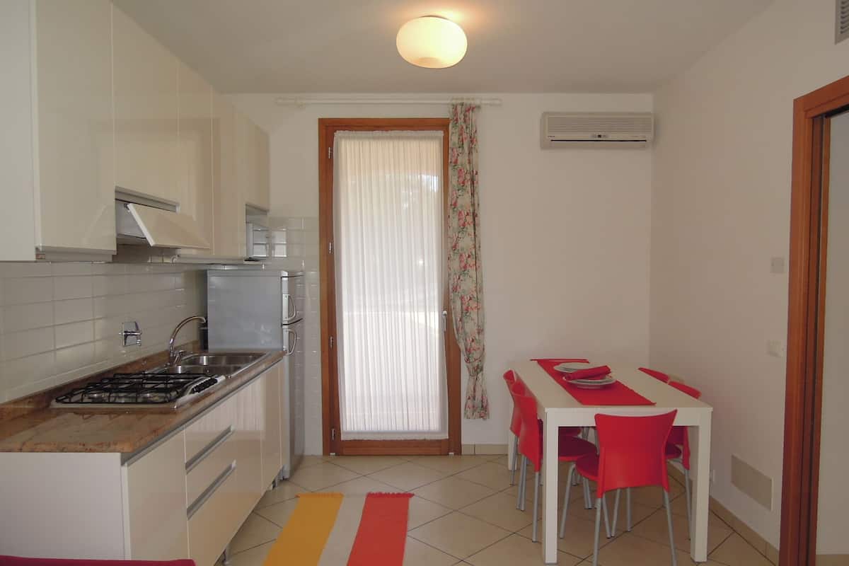 A compact kitchen with a gas stove, sink, and white cabinets. There is a dining area with a table and red chairs.