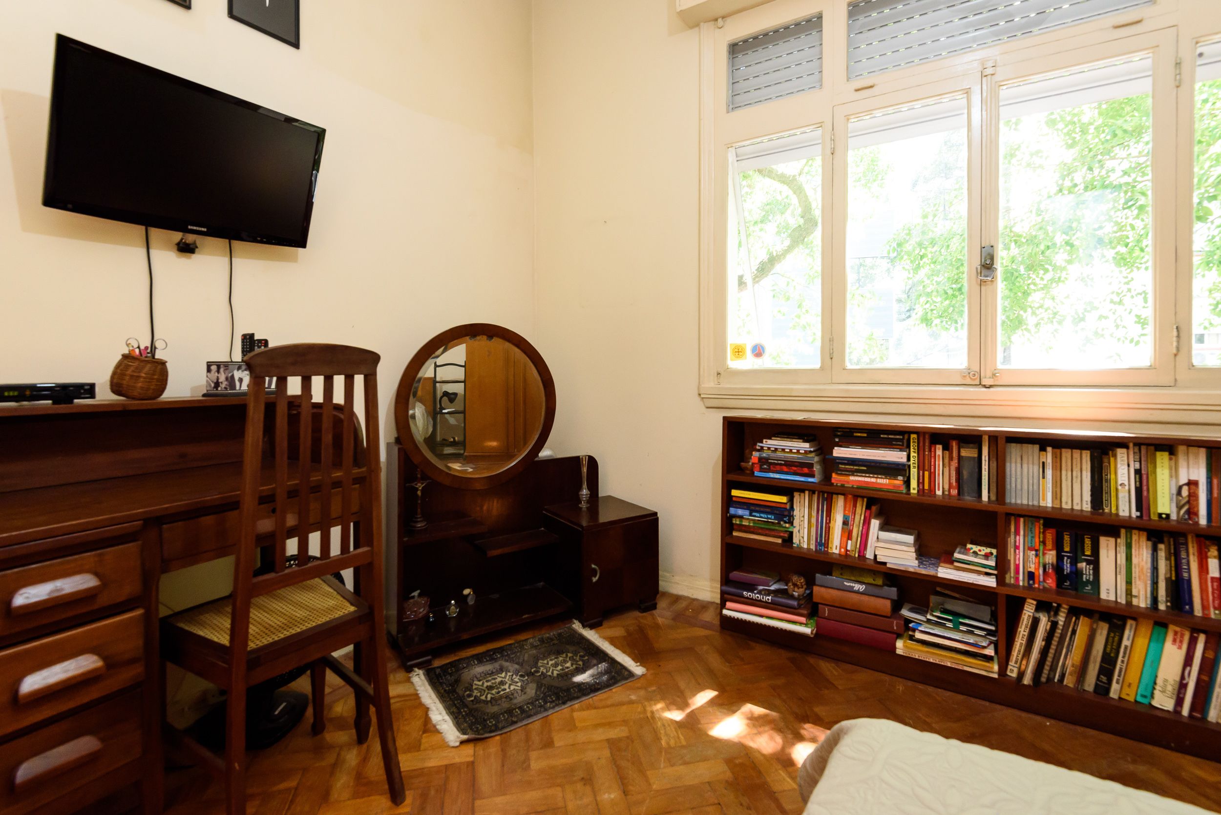 A room with a wooden desk, a chair, a bookshelf, a television, and a window with a view of trees.