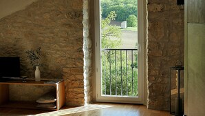 A room with a stone wall, a wooden floor, a bed, a TV, a vase with plants, and a balcony with a view of greenery. - La Bastide (Lamillarie)