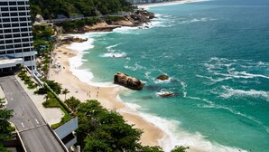 A coastal cityscape with a beach, a large rock formation, and a modern building with a road leading to the beach. - Omar do Rio - Apartamento V902 (Rio de Janeiro)
