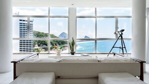 A modern living room with a large window overlooking a beach and cityscape. - Omar do Rio - Apartamento V902 (Rio de Janeiro)