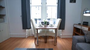 A modern living room with a dining table, two chairs, a grey sofa, a clock, and a window with curtains.