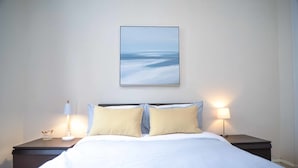 A neatly made bed with white linens and two pillows, flanked by two bedside tables with lamps, one of which has a small decorative item on it.