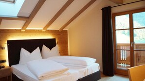 A bedroom with a wooden headboard, a bed with white linens, a nightstand, and a large window with a view of mountains. - Gasthof Laurin (Tires)