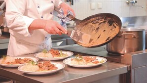 A chef in a white uniform with a hat, preparing food in a kitchen with stainless steel counters and various utensils hanging on the wall. - Gasthof Laurin (Tires)