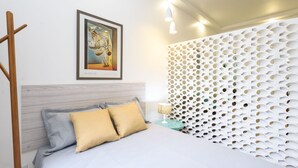 A neatly arranged bedroom with a bed, pillows, a bedside table with a lamp, a framed picture on the wall, and a decorative white perforated panel.