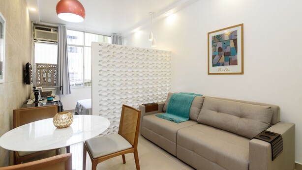 A modern living room with a beige sofa, a round dining table with chairs, a decorative vase, and a wall-mounted artwork.