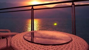 Una mesa redonda de vidrio con una silla tejida, barandilla de balcón y vista al atardecer sobre el agua.