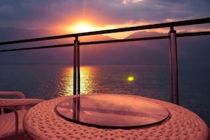 A round glass table with a woven chair, a balcony railing, and a sunset view over water. - Muhai Resort Apartment (Huidong)