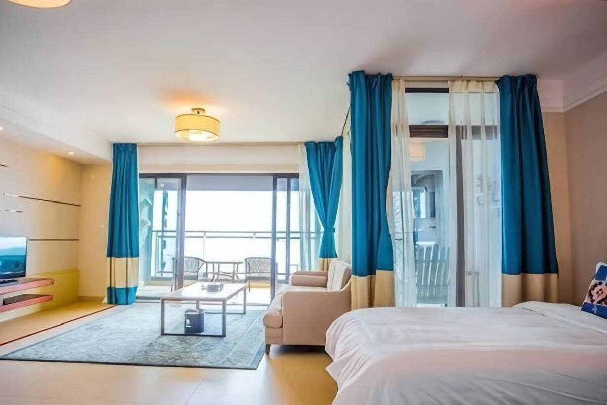 Quarto de hotel com cama, sofá, uma mesinha e televisão. Possui varanda com cortinas azuis.