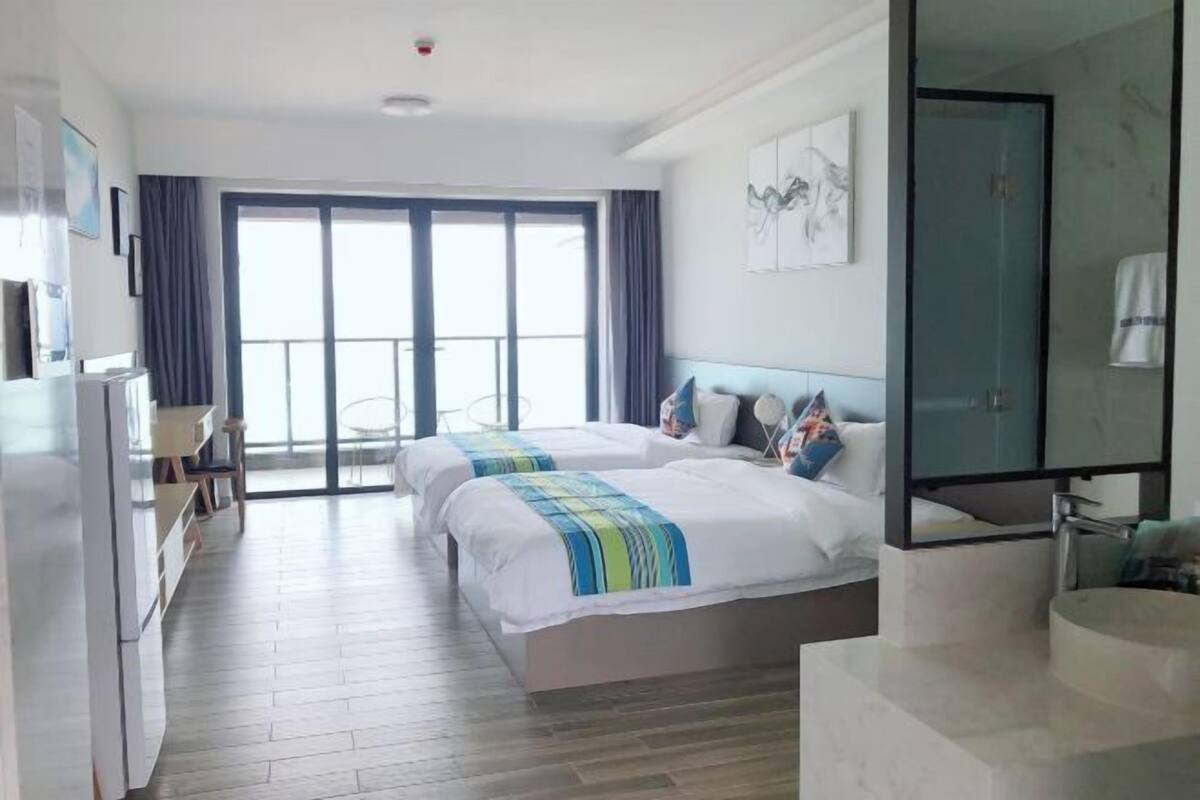 Quarto de hotel com duas camas, uma escrivaninha, uma cadeira, uma janela grande e banheiro com chuveiro de vidro.