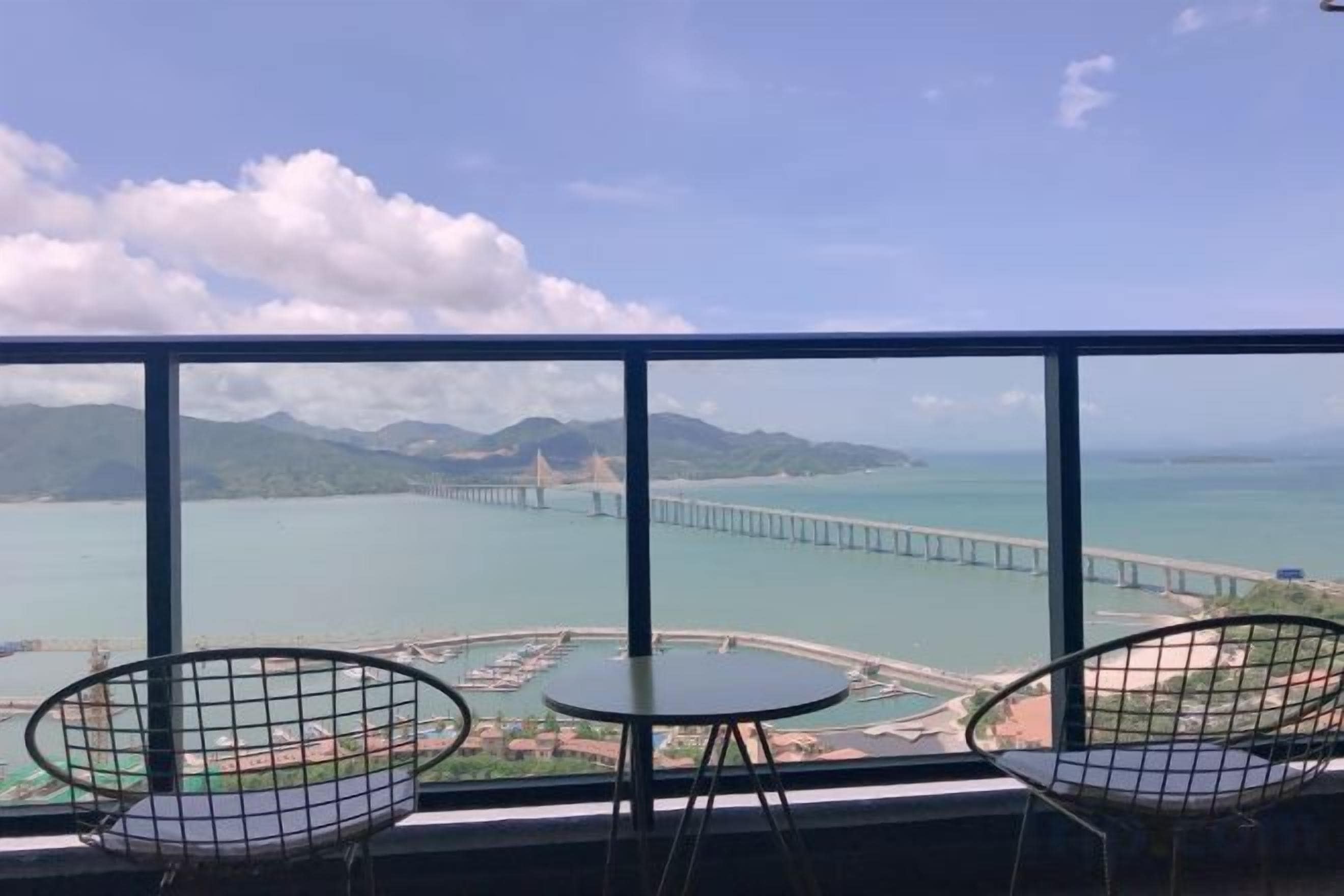 Uma varanda com duas cadeiras e uma mesa, com vista para um corpo de água, uma ponte e montanhas ao longe.