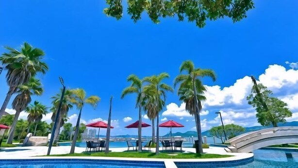 Zona de piscina con palmeras, sombrillas rojas y un puente.