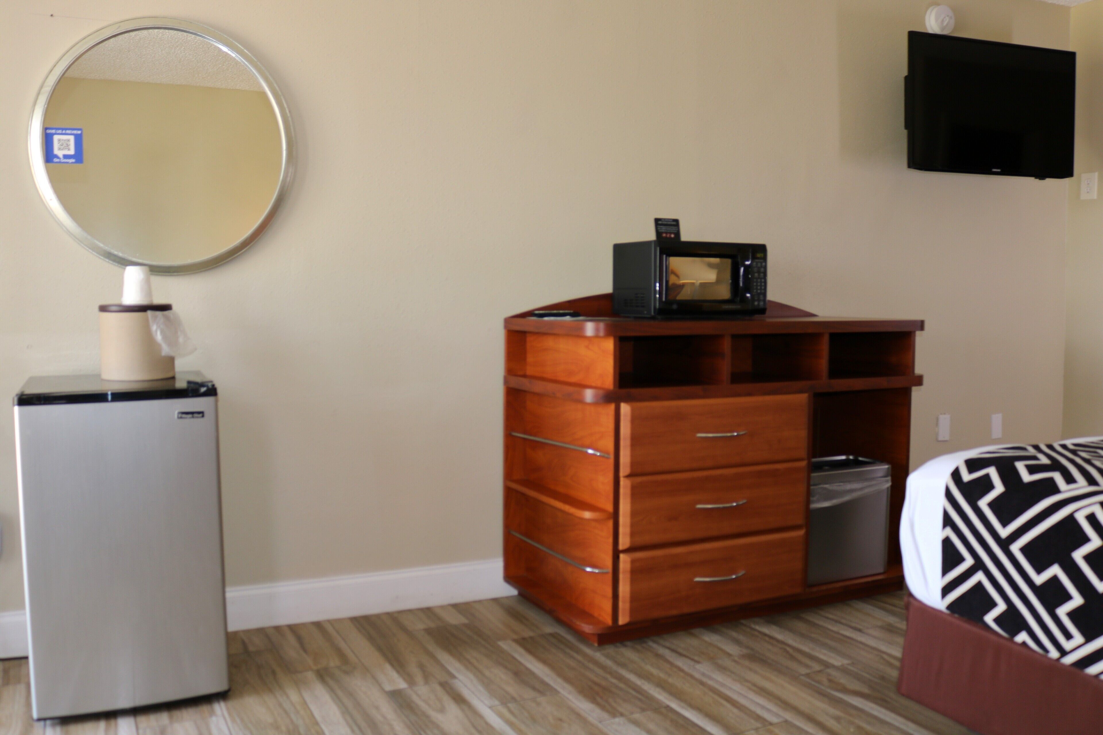 A compact room with a wooden dresser, a small refrigerator, a flat-screen TV mounted on the wall, and a bed with a patterned bedspread.