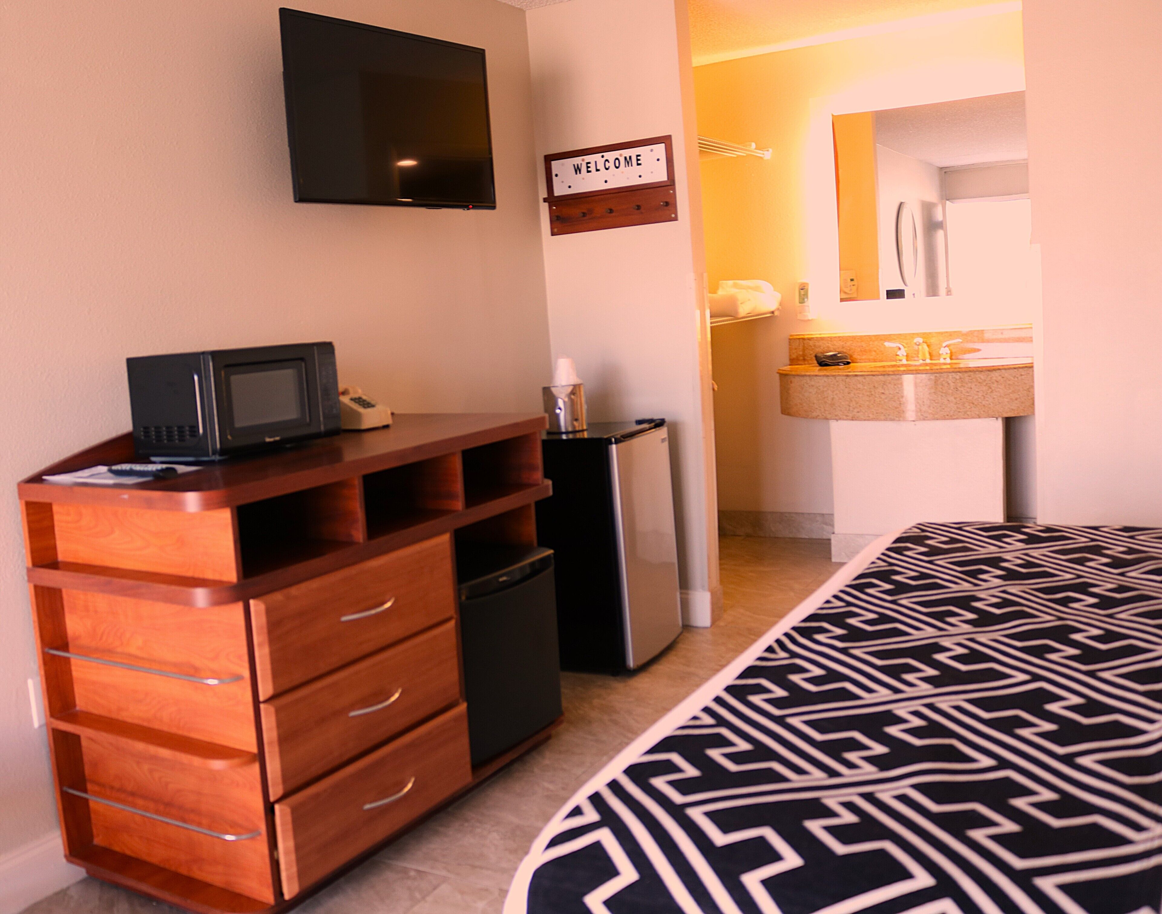 A compact hotel room with a wooden dresser, a microwave, a refrigerator, a bed with a patterned bedspread, a television mounted on the wall, and a welcome sign.