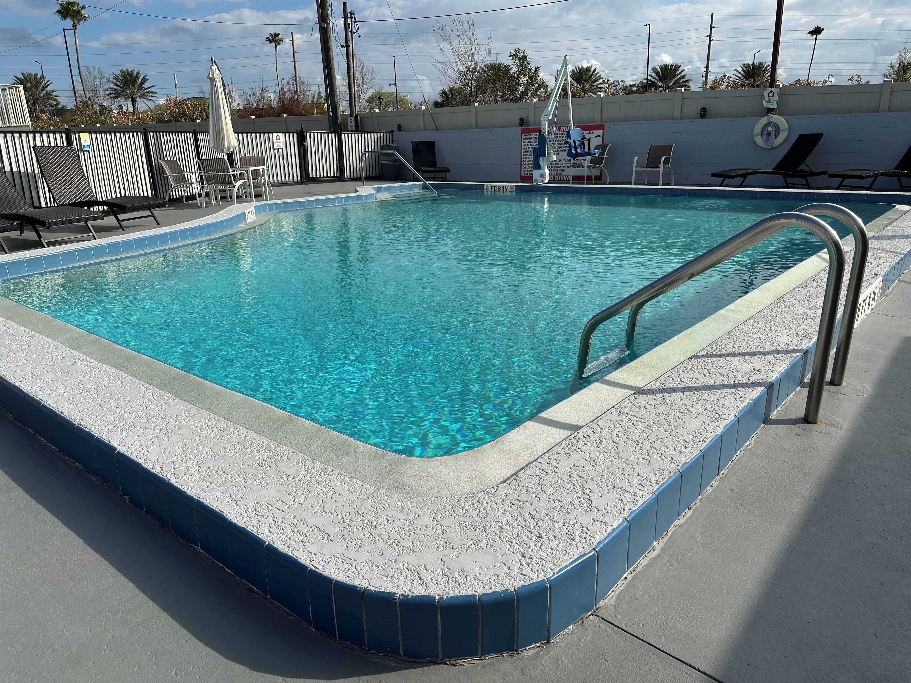 A rectangular swimming pool with a blue tiled edge, a metal handrail, and lounge chairs along the side.