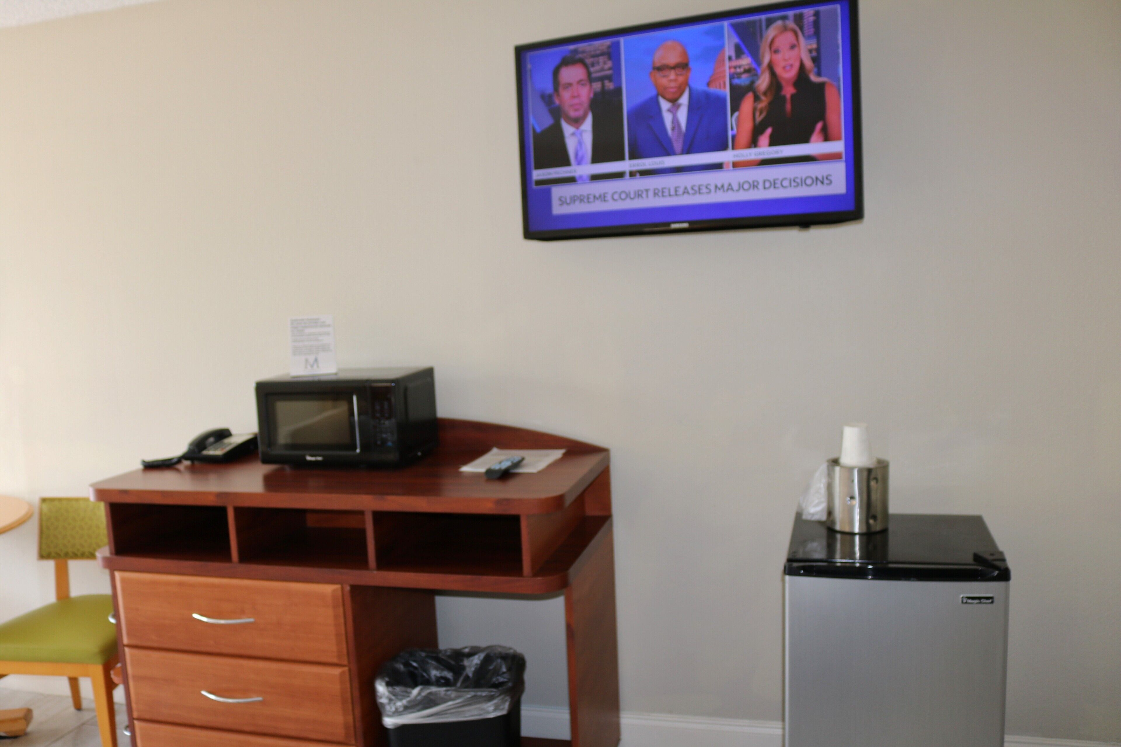 A hotel room with a television displaying a news program, a wooden desk with a telephone, a trash bin, and a small refrigerator.