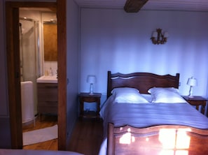 A bedroom with a wooden bed, two bedside tables with lamps, a nightstand, a wooden wardrobe, and a bathroom visible through an open door. - La Ferme Des Vignes (Hotot-en-Auge)