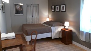A bedroom with a bed, two bedside lamps, a chair, a table, and a window with curtains. - La Ferme Des Vignes (Hotot-en-Auge)