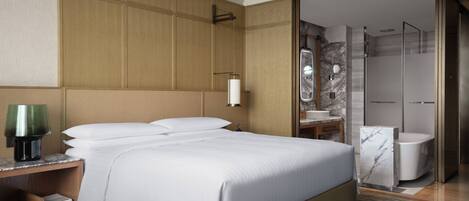 Una habitación de hotel moderna con una cama grande, una mesita de noche con lámpara, un baño con ducha de mármol y un vestidor.