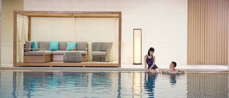 A modern indoor pool area with a seating area featuring a sofa and cushions, a large framed picture, and a wooden ceiling structure.