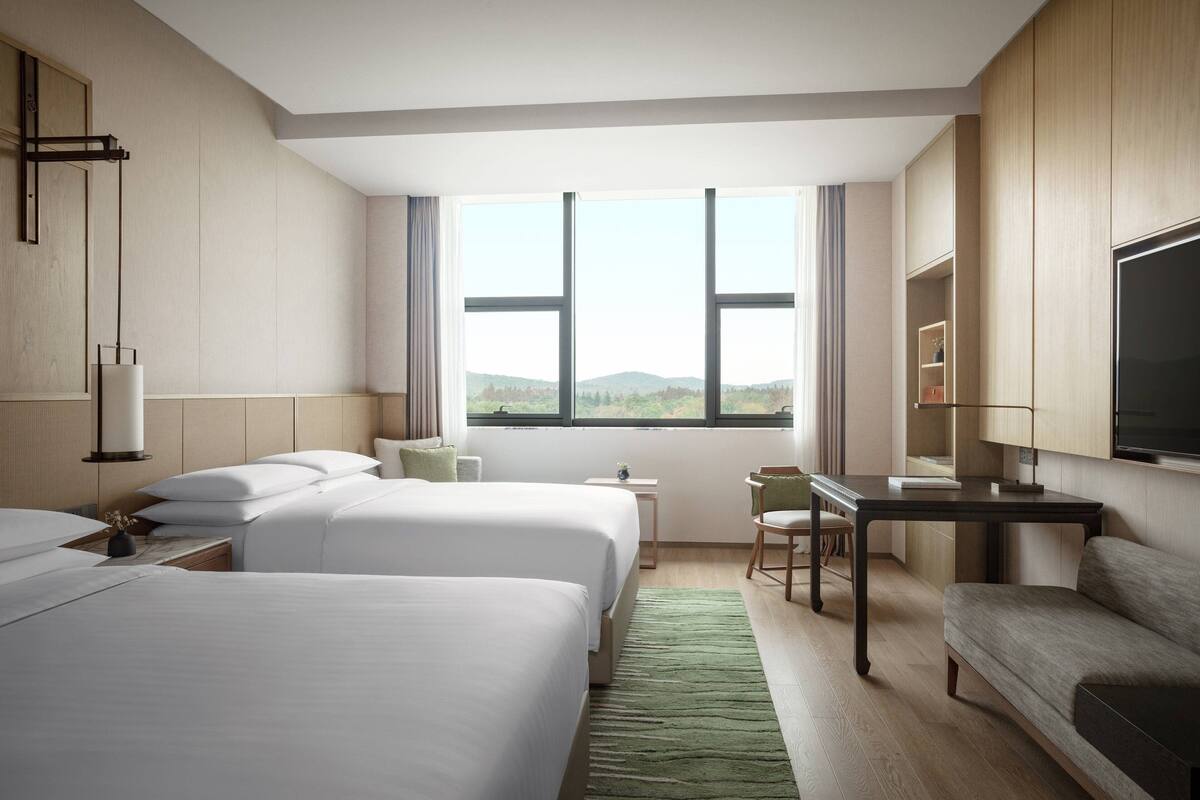 A hotel room with two beds, a desk, a chair, a TV, and a view of the outdoors.