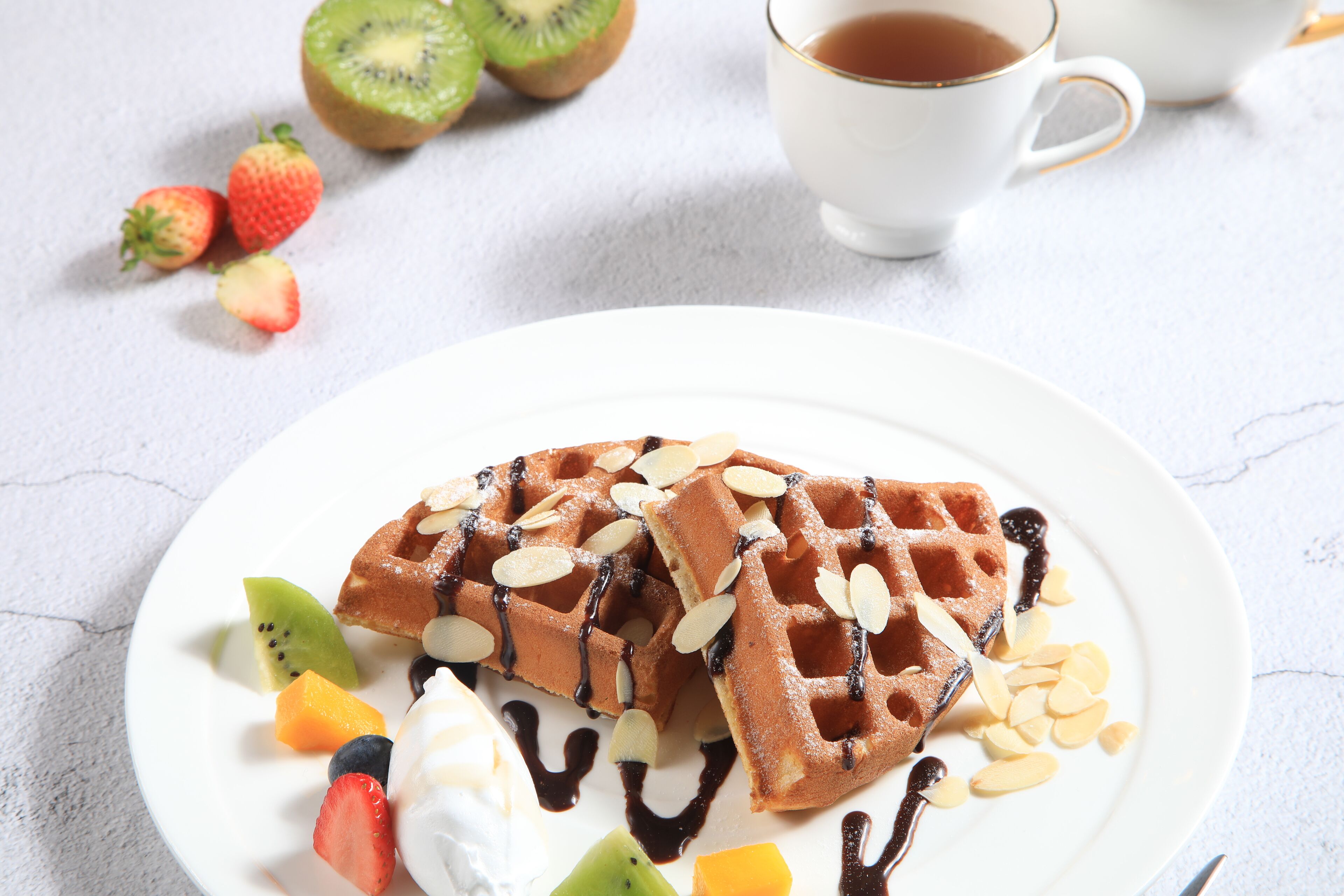 A plate of waffles with fruit and almonds, a cup of tea, and a few strawberries and kiwis on a table.