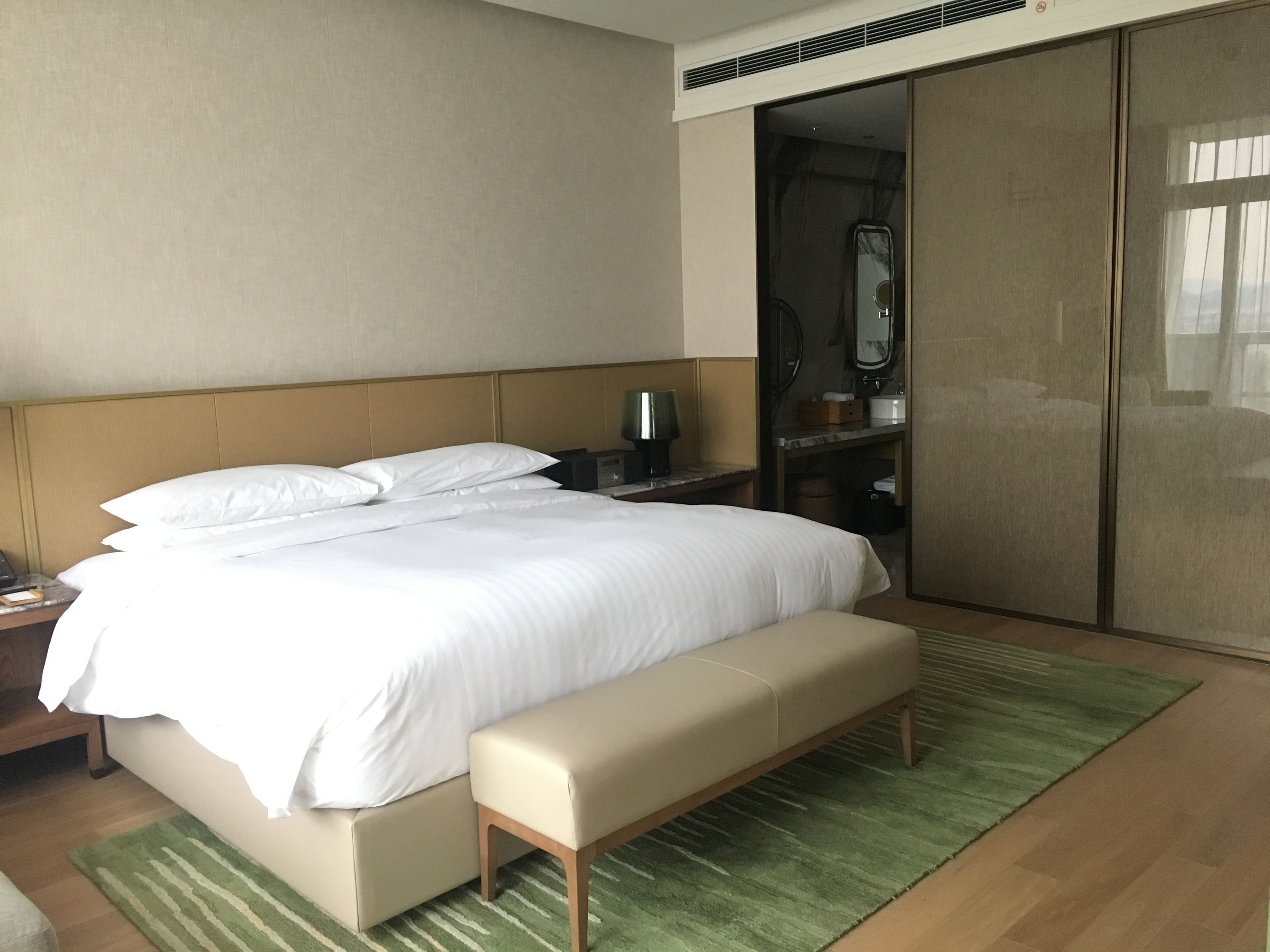 A hotel room with a large bed, a bench, a nightstand, a mirror, and a wardrobe.