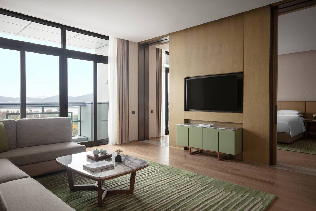A modern hotel room with a large window, a flat-screen TV, a green minibar, and a sofa.