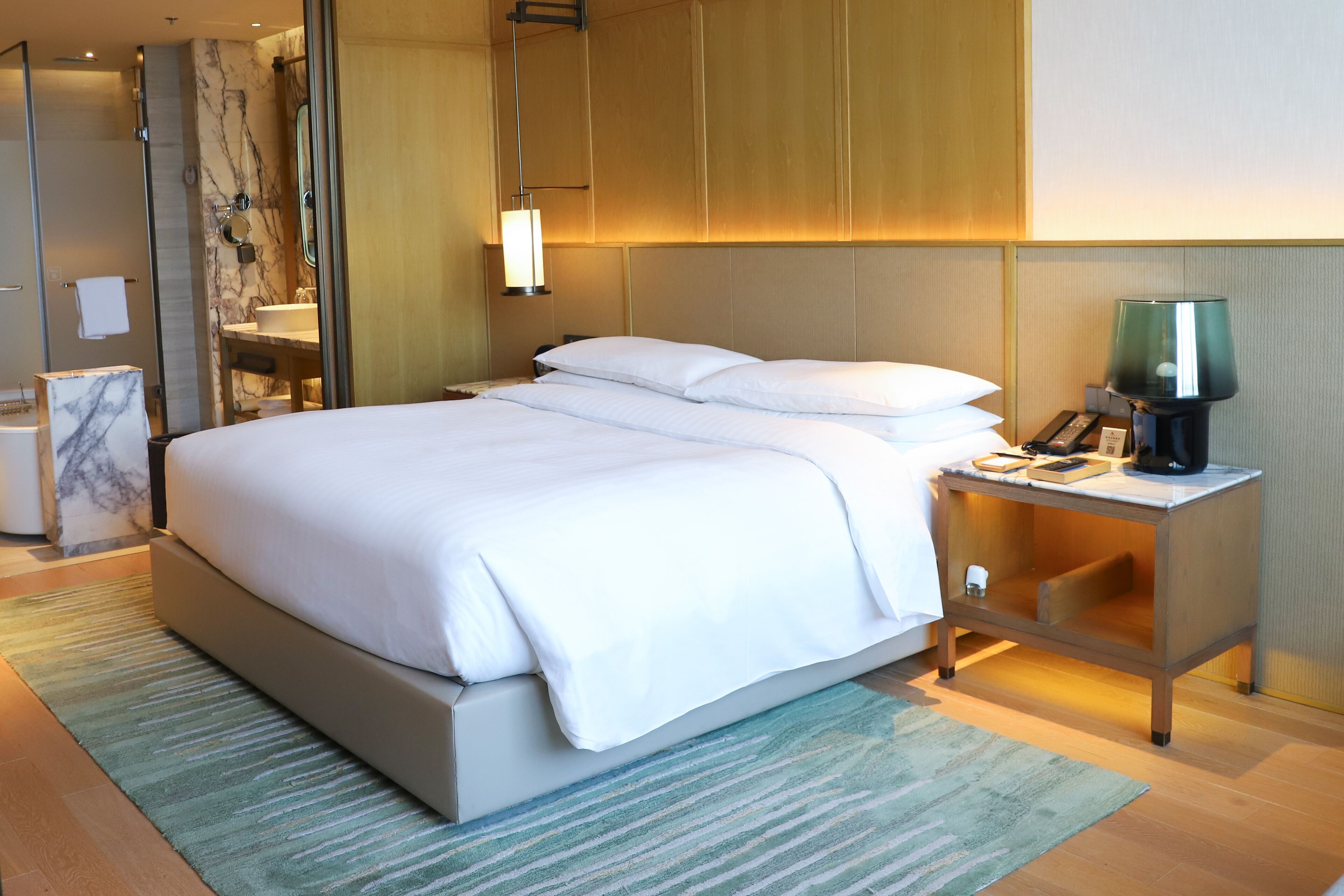 Une chambre d’hôtel moderne avec un grand lit, une table de chevet avec une lampe, une commode surmontée d’un plateau en marbre et un espace douche doté d’une porte en verre.