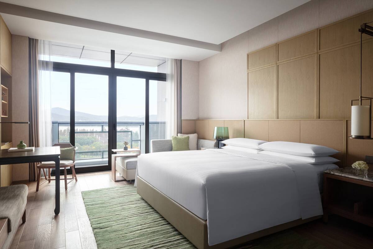 A modern hotel room with a large bed, a desk, a chair, and a view of the cityscape through large windows.