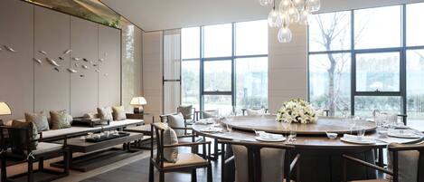 A modern dining area with a round table set for six, surrounded by six chairs. There is a sofa set with cushions and a coffee table in the background.