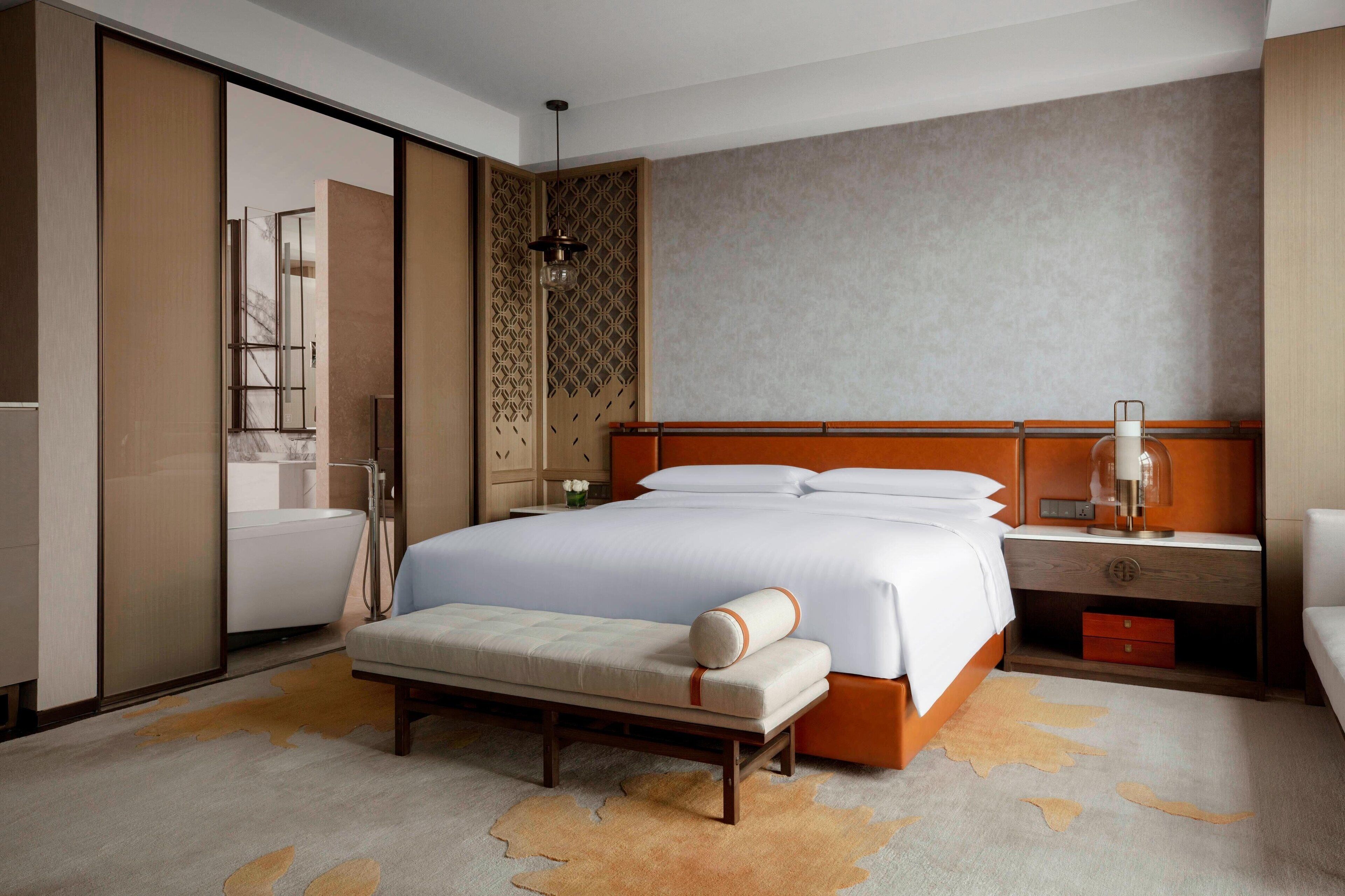 A modern hotel room with a large bed, a bench, a nightstand, and a bathroom visible through a sliding door.
