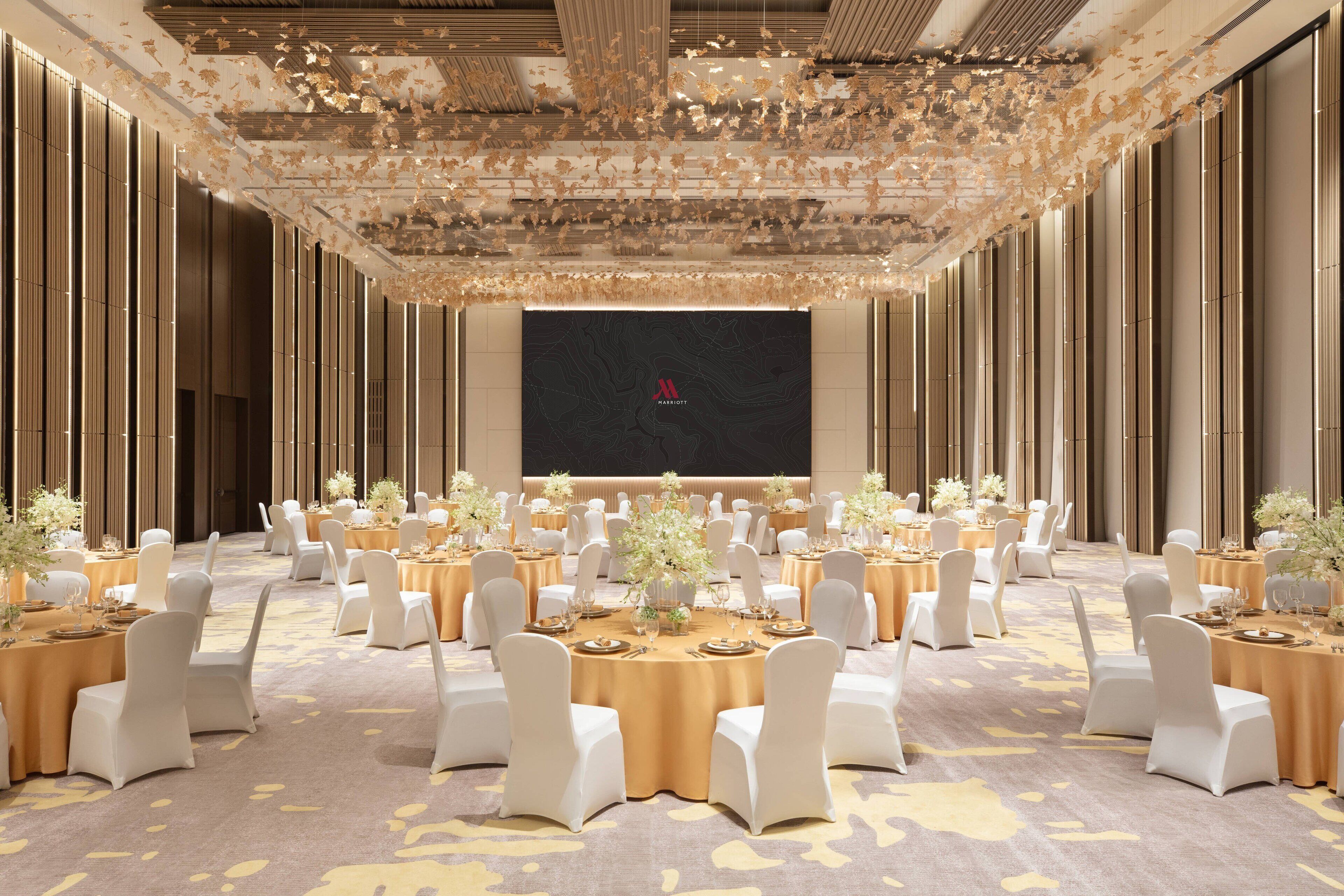 Un amplio salón de banquetes con mesas redondas, sillas blancas y centros de mesa florales.