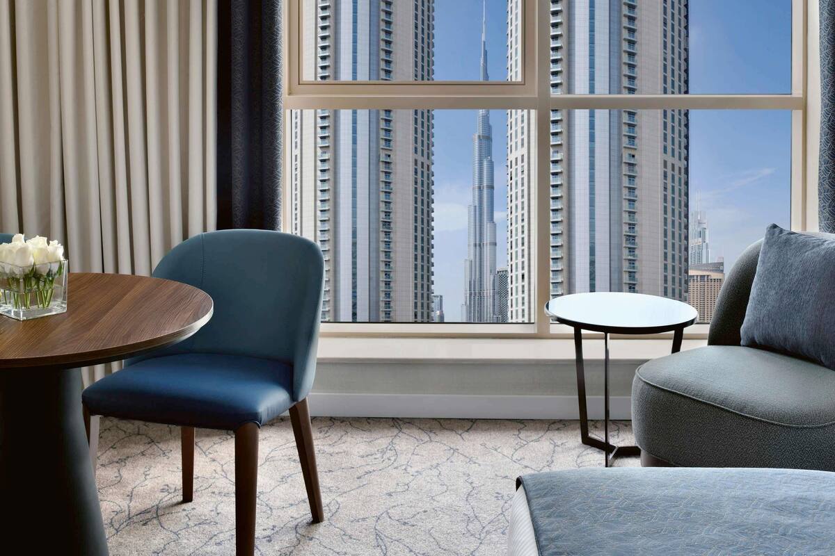 A modern room with a view of tall buildings, a blue chair, a small round table with a vase of flowers, and a sofa.