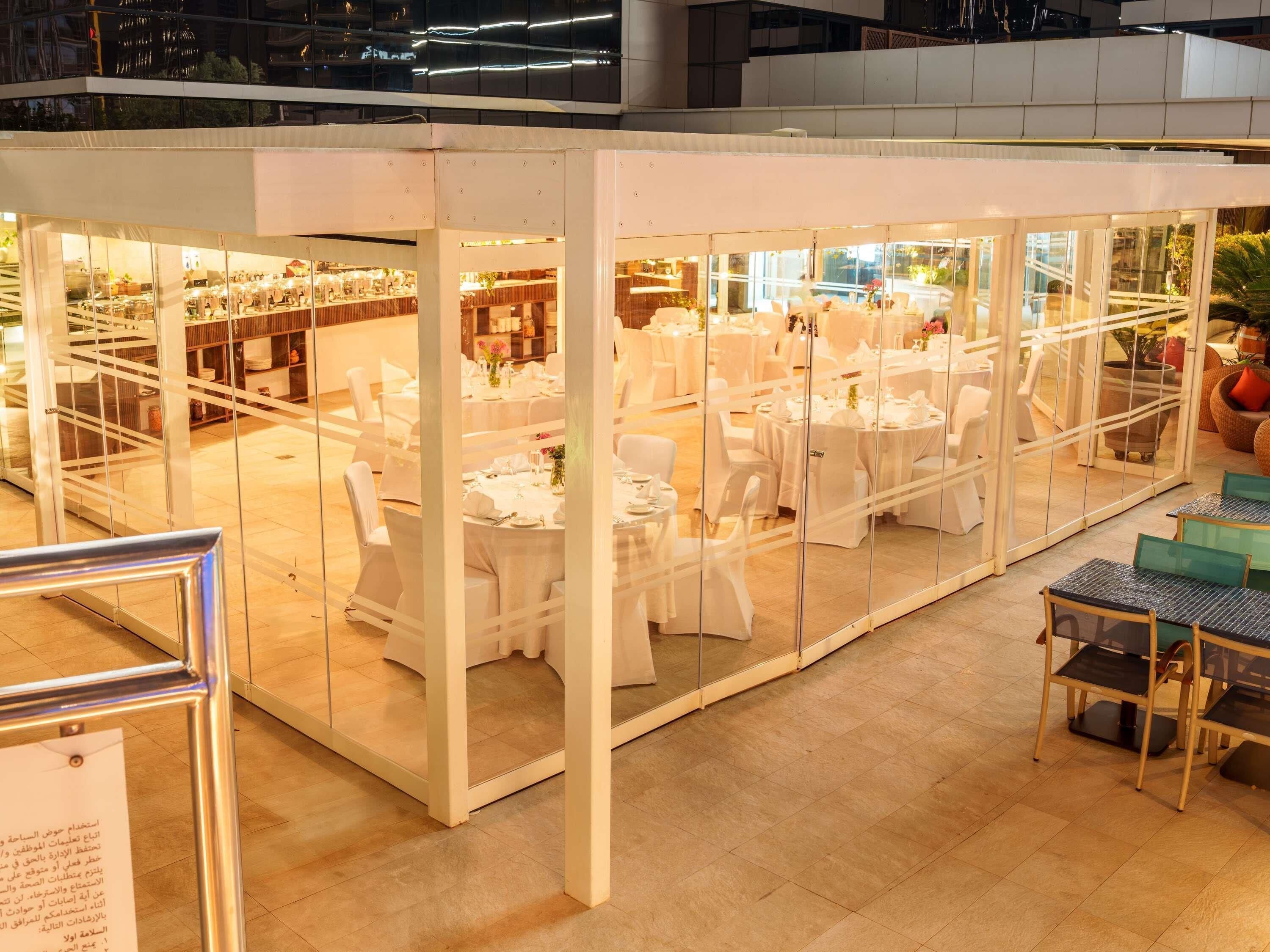 A glass-enclosed dining area with tables set for a meal, surrounded by white chairs.