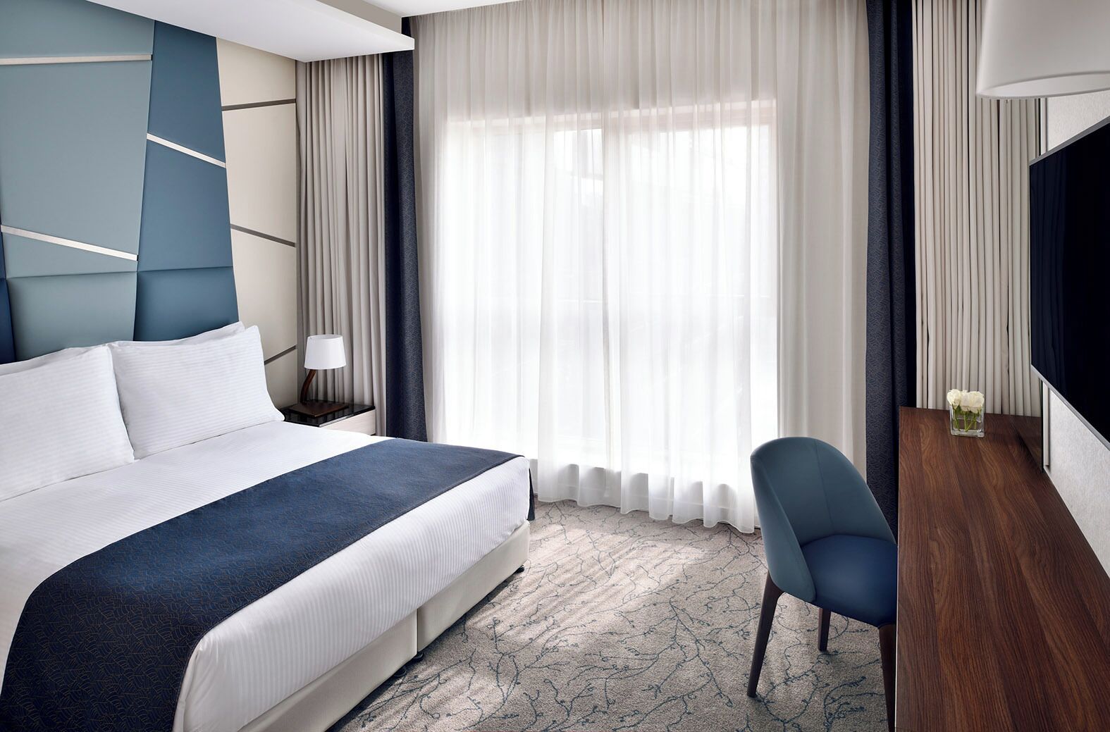 A modern hotel room with a large bed, a desk, a chair, and a TV.