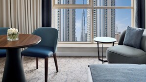 A modern hotel room with a large window offering a view of high-rise buildings. There is a round wooden table with a vase of white flowers, a blue armchair, and a grey sofa. - Mövenpick Hotel Apartments Downtown Dubai (Dubai)