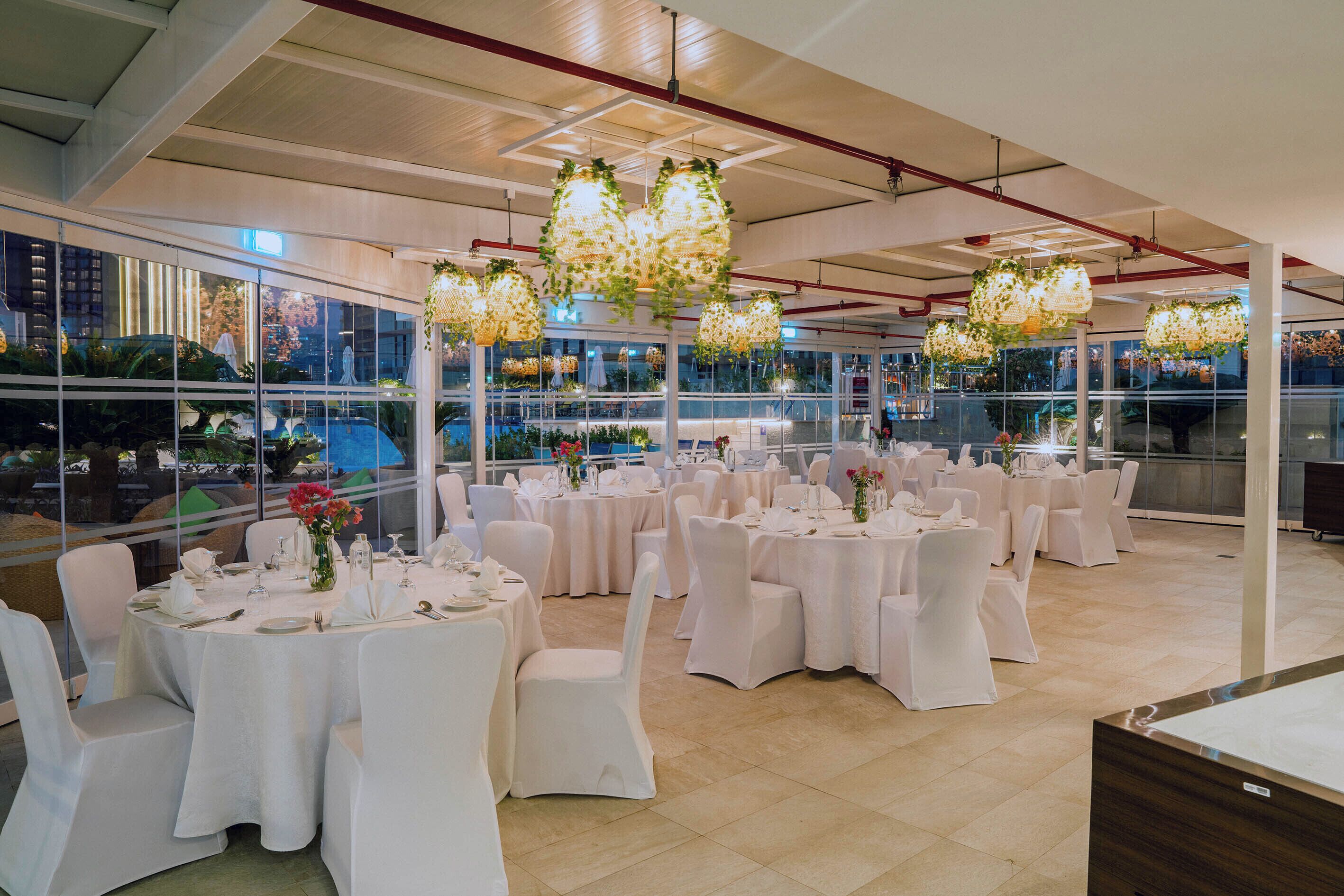 A spacious indoor venue with round tables set for dining, large windows, and hanging floral light fixtures.