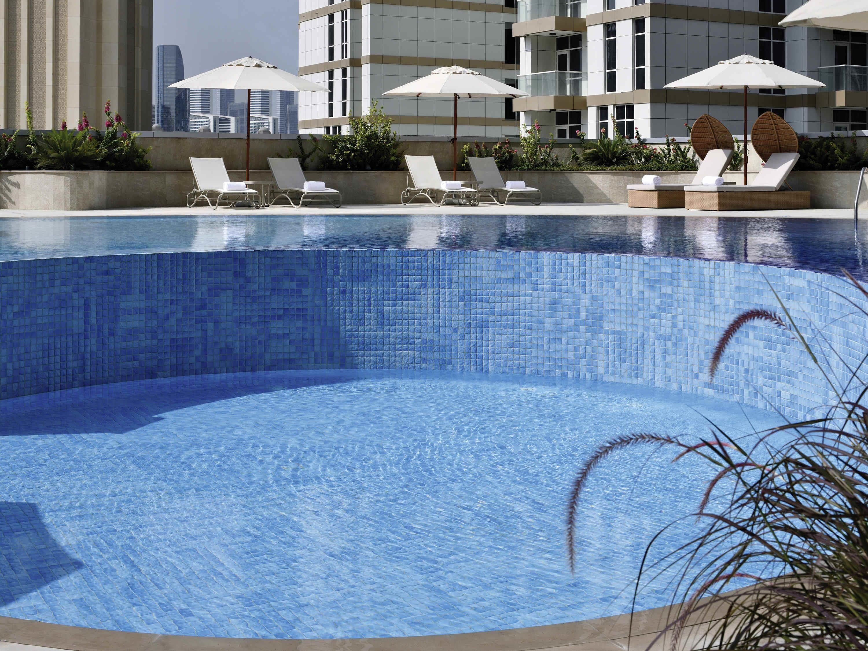 A swimming pool with lounge chairs and umbrellas, surrounded by a modern building with a tiled facade.