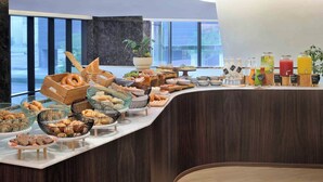 Un buffet proposant une variété de plats et de boissons, une grande plante en pot et une vue sur l’extérieur grâce à de grandes fenêtres.