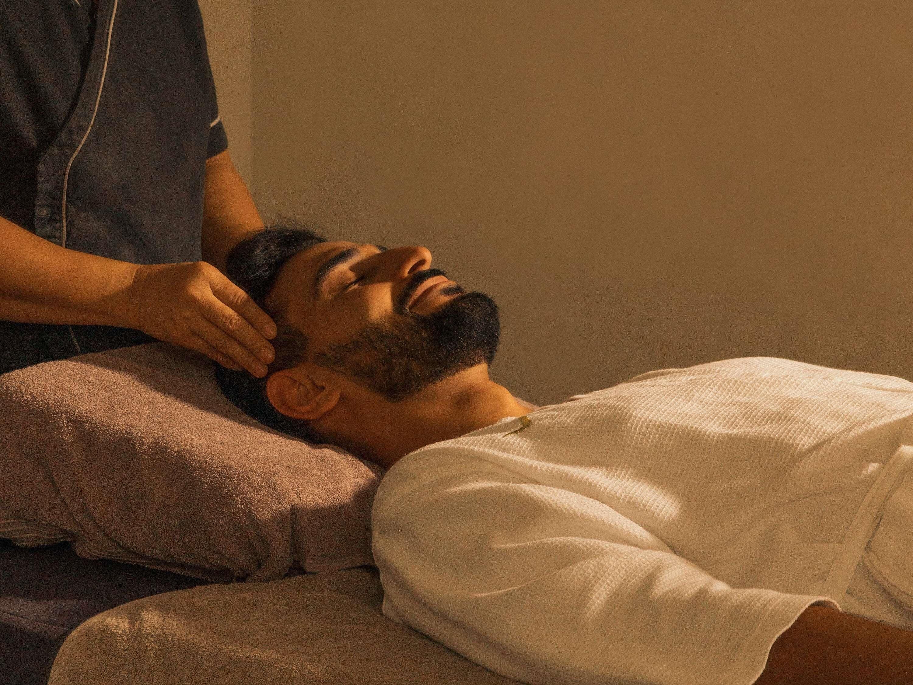 A person lying on a bed with a pillow, wearing a white shirt, in a room with a plain wall.