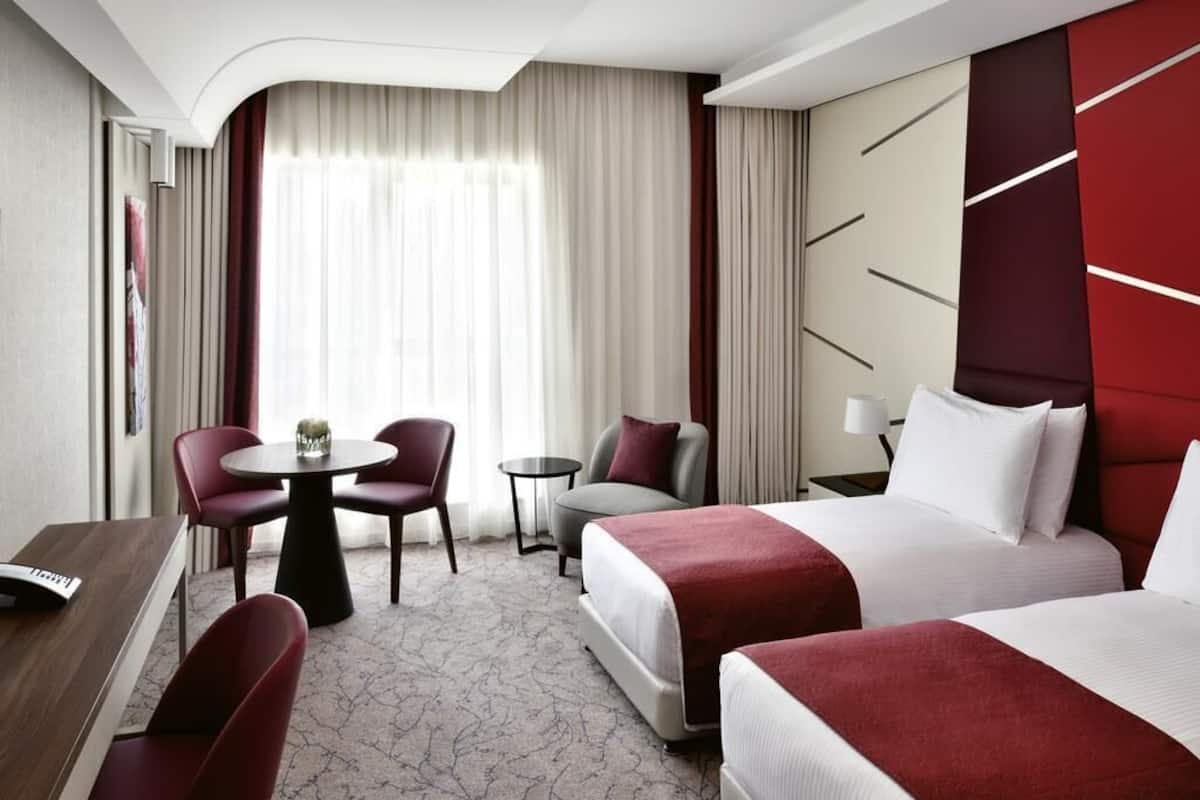 Une chambre dâhĂŽtel avec deux lits, un bureau, une chaise, une petite table et une fenĂȘtre avec des rideaux.