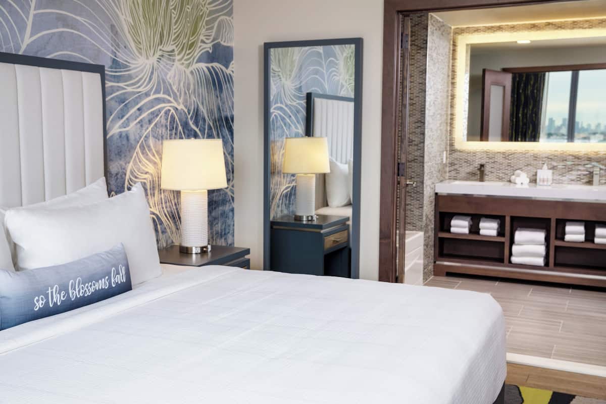 Habitación de hotel con cama, dos lámparas de noche, un espejo grande y baño con lavabo y toallero.