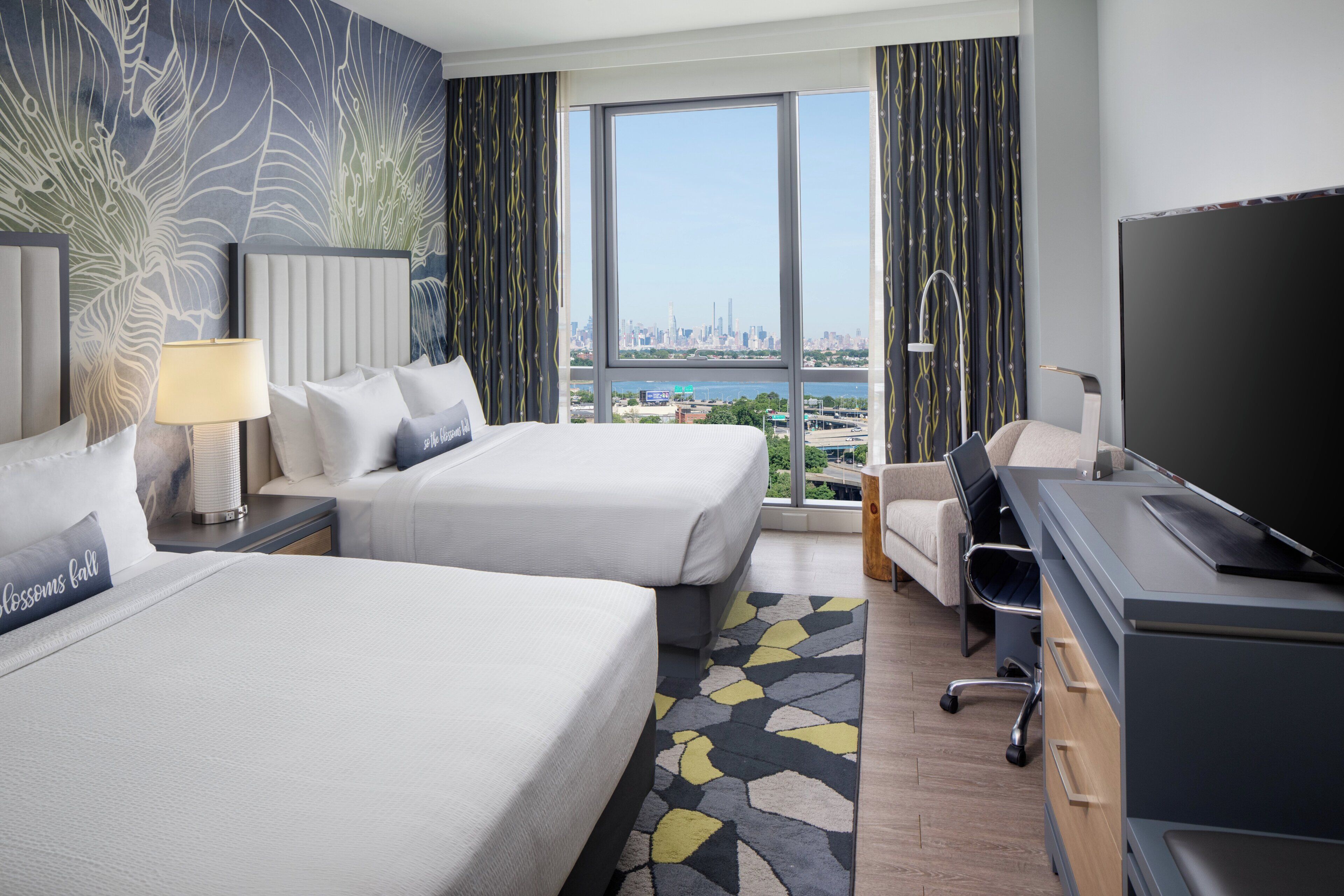 A hotel room with two beds, a desk, a TV, and a view of a cityscape.